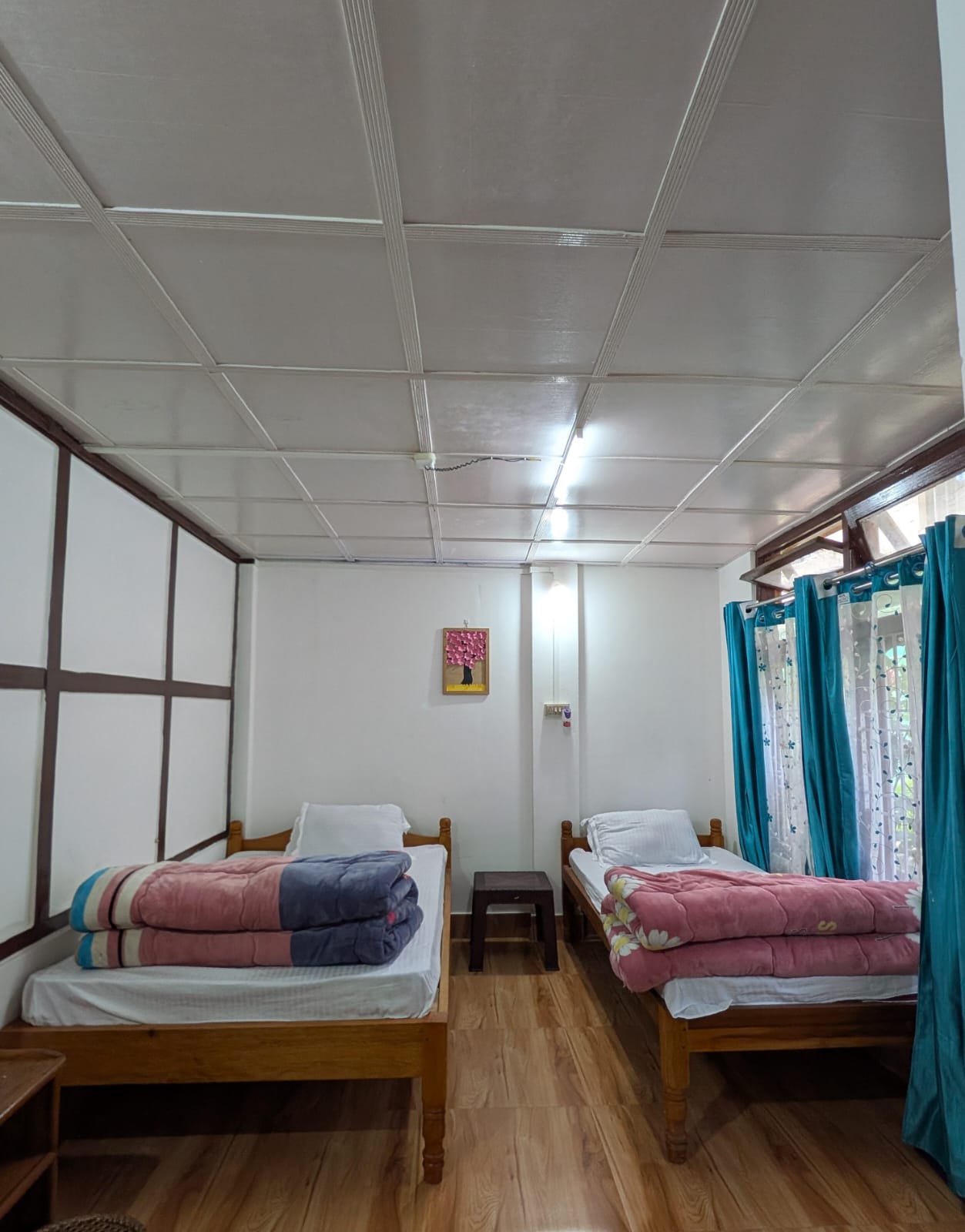 Two wooden beds with folded quilts, teal curtains, and a small picture of a pink tree
